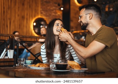 Smiling man in casual attire feeds his partner a pastry in a cozy restaurant setting, indicative of a cheerful dining moment. - Powered by Shutterstock