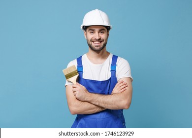 Smiling man in coveralls protective helmet hardhat hold paint brush isolated on blue wall background. Instruments accessories for renovation apartment room. Repair home concept. Holding hands crossed