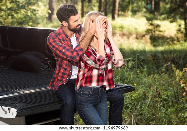 smiling man covering eyes of girlfriend\
from behind while sitting on car trunk outdoors\
