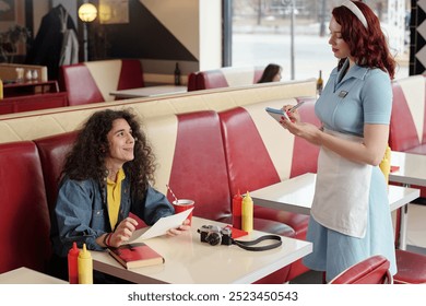 Smiling man with curly hair sitting at diner booth, interacting with waitress writing order on notepad, vintage camera and a notebook on table, ketchup and mustard dispensers - Powered by Shutterstock