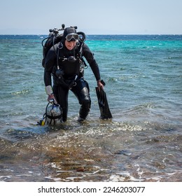 A smiling man diver in scuba gear with one cylinder in his right hand, fins in his left hand and twin cylinders comes out of the sea, after finishing a technical dive - Powered by Shutterstock