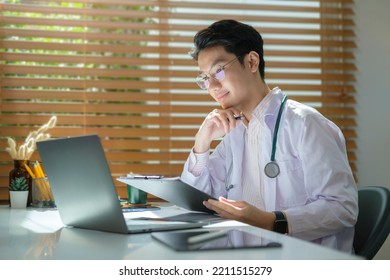 Smiling Man Doctor Wearing White Uniform Reading Electronic Medical Records On Laptop Computer.