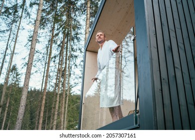 Smiling Man Dressed White Bathrobe Standing On Forest House Balcony And Enjoying Fresh Air With Nature Forest Pine Trees View. Outside Scandinavian Interior Design House. Living In Wild Concept Image