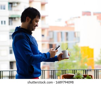 Smiling man drinking coffee on the balcony of his house and looking at his smartphone. Coronavirus confinement