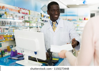 Smiling Man Druggist Working Behind Counter In Pharmacy, Taking Prescriptions From Customer