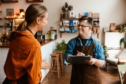 Smiling Man Employee With Down Syndrome Wearing Apron Writing Down Notes In Clipboard While Young Smiling Woman Instructing Him In His Waiter Duties