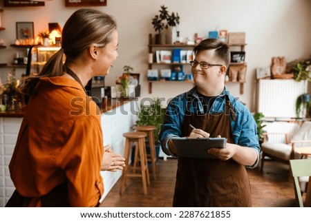 Smiling man employee with down syndrome wearing apron writing down notes in clipboard while young smiling woman instructing him in his waiter duties