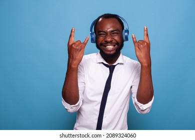 Smiling Man Full Of Energy Doing Rock And Roll Hand Sign While Listening To Music In Wireless Headphones. Business Man Acting Rebel Doing Hand Sign While Listening To Loud Music In Earphones.