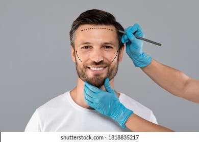 Smiling man getting pencil marks on face skin for cosmetic surgery, ready for operation. Aesthetic Cosmetology concept. Plastic surgeon applying marks on male face before surgery, grey background