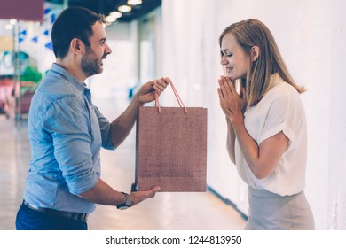 Smiling Man Giving Gift In Paper Bag To Excited Wife In Mall With Glass Walls In Background. Gift Concept. Side View.