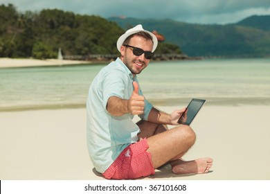 smiling man giving thumbs up while sitting on the beach and reading on an ipad