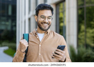 Smiling man with glasses holding smartphone outdoors near modern office building. Casual attire, backpack, and glasses emphasize relaxed and tech-savvy persona. Captures urban lifestyle - Powered by Shutterstock