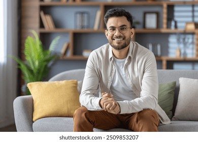 Smiling man with glasses relaxing on sofa in modern living room. Casual style with beige shirt and white t-shirt. Bright, cozy atmosphere reflects warmth and tranquility. - Powered by Shutterstock