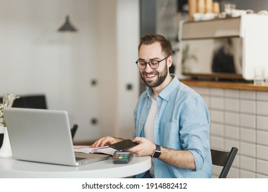 Smiling Man In Glasses Sit At Table In Coffee Shop Cafe Restaurant Indoors Working Studying On Laptop Computer Pay Off With Cell Phone Bank Payment Terminal. Freelance Mobile Office Business Concept