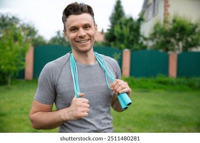 Smiling man in a gray athletic shirt stands outdoors, holding a light blue jump rope draped around his neck. The background features a green fence and trees, emphasizing a cheerful, active lifestyle - Powered by Shutterstock