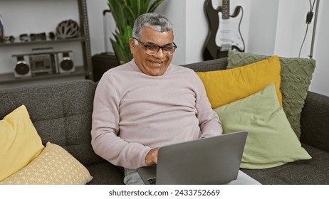 A smiling man with grey hair sits comfortably on a grey couch with colorful pillows while working on a laptop in a cozy living room. - Powered by Shutterstock