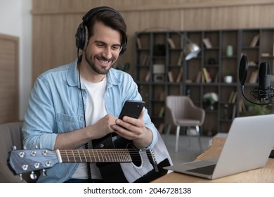 Smiling man in headphones using smartphone, sitting at desk with guitar, happy musician artist reading good comments in social networks, received good news, great offer, working in home studio