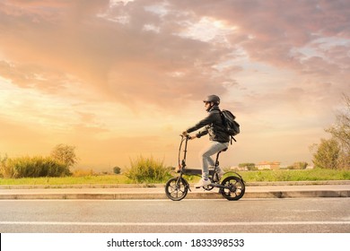 Smiling Man With Helmet And His Electric Bicycle At Sunset. Commuter And New Transportation Concept. Green Energy. Caucasian Man Drives His Foldable E Bike To Go Work Or In Leisure