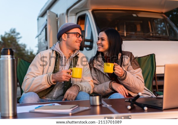 Smiling man holding\
cup with tea and squints against the bright sun near his woman.\
Happy family sitting at the nature and drinking hot tea from cups\
at camp fire in cozy\
forest