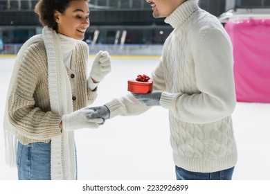 Smiling man holding heart shaped box and hand of african american girlfriend on ice rink  - Shutterstock ID 2232926899
