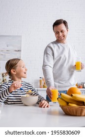 smiling man holding orange juice near daughter eating corn flakes for breakfast
