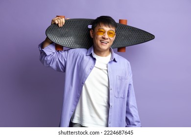 Smiling Man Holding Skateboard. Asian Guy Holding Longboard on Shoulder and Smiling while Posing Isolated on Violet Background 