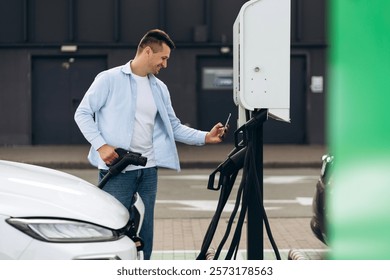 Smiling man holding the type2 EV charger plug handle near electric car while paying by smartphone outdoors. Alternative clean and sustainable energy concept