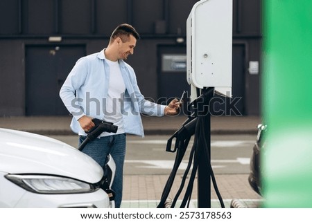 Smiling man holding the type2 EV charger plug handle near electric car while paying by smartphone outdoors. Alternative clean and sustainable energy concept