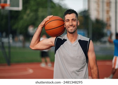 Smiling man holds a basketball on an outdoor court with players in background, depicting sport, basket, and ball elements - Powered by Shutterstock