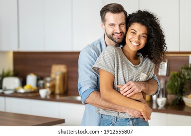 Smiling man hugging from behind charming African American woman, two people standing and joyfully looking at camera. Young international couple happily spending time in cozy modern kitchen at home. - Shutterstock ID 1912555807