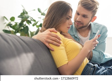 Smiling man hugging depended girlfriend with smartphone on couch - Shutterstock ID 1712860972
