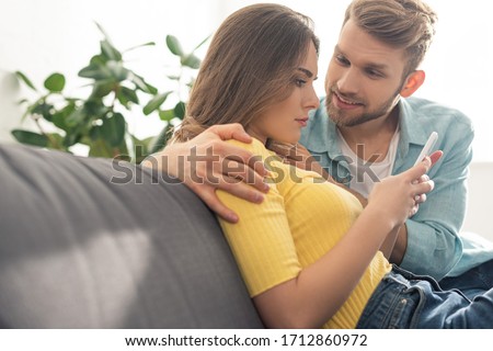Smiling man hugging depended girlfriend with smartphone on couch