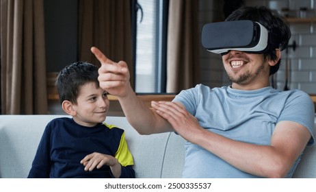 Smiling man immersed in a virtual reality experience with a VR headset while sits on sofa with young boy. Modern family life, the immersive and interactive of VR technology in a cozy home - Powered by Shutterstock