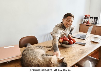 Smiling man interacts with his cat while working at a wooden table filled with fresh fruit. - Powered by Shutterstock