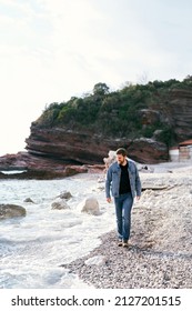 Smiling Man In Jeans And Denim Jacket Walking Along The Pebble Beach Looking At The Water Against The Background Of Green Rocks