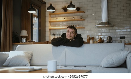 Smiling man leaning over the back of the sofa puts on the smartphone on sofa and ponder deep in thoughts dreaming at home. - Powered by Shutterstock