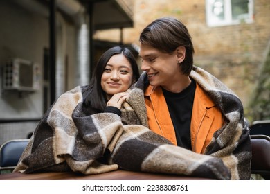 Smiling man looking at asian girlfriend in blanket in outdoor cafe - Shutterstock ID 2238308761