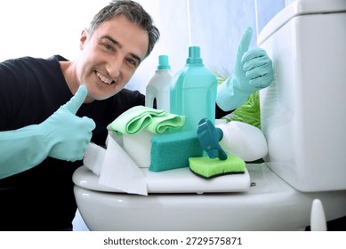 Smiling man looking at camera making ok gesture with hands wearing rubber gloves with various cleaning products on toilet bowl in bathroom with blue tiles.