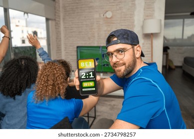 Smiling man looking at camera while showing a smartphone screen with a sports betting website as football soccer plays at home. Online wagering, match prediction, modern gambling, digital sports entertainment