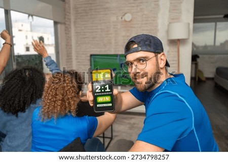 Smiling man looking at camera while showing a smartphone screen with a sports betting website as football soccer plays at home. Online wagering, match prediction, modern gambling, digital sports entertainment