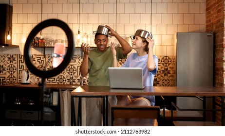 Smiling Man Looking On Offended Girl During Making Video In Prank Or Challenge On Smartphone For Social Networks. Multiracial Blogger Couple Wearing Saucepan On Heads At Table On Home Kitchen