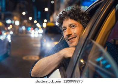 Smiling man looking outside from car window at night. Handsome business man enjoying taxi ride viewing the cityscape. Happy man traveling by taxi during vacation.