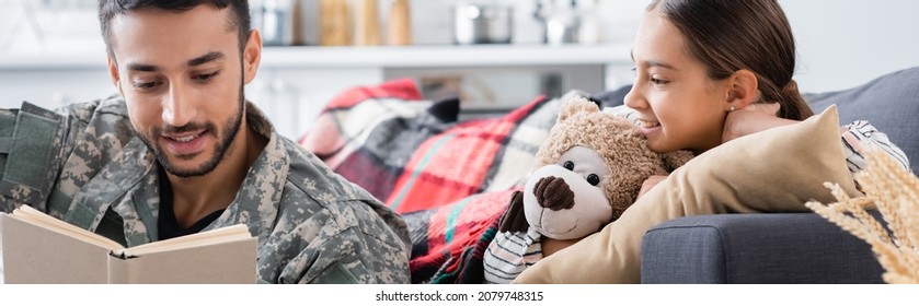 Smiling Man In Military Uniform Reading Book Near Child With Teddy Bear On Couch, Banner