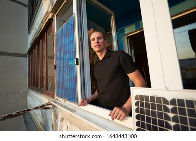 Smiling man on the balcony of a multi-storey building equipped with a solar panel, against a background of a residential area of the city with clear blue sky on a warm evening - Powered by Shutterstock