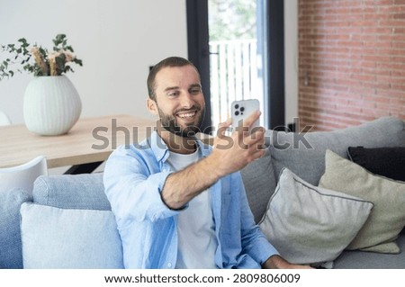 Smiling man on a couch taking a selfie video call with his smartphone in a bright living room, relaxed and casual at home, enjoying remote communication and staying connected with friends or family