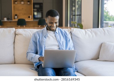 Smiling man on couch using laptop work, relaxed home interior scene conveying focus and happiness, casual professional in blue shirt, remote work lifestyle, modern livingroom comfort and productive atmosphere.