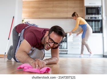 Smiling Man On His Knees Polishes Floor With A Cloth While Woman In Background Putts The Dishes In Dishwasher.