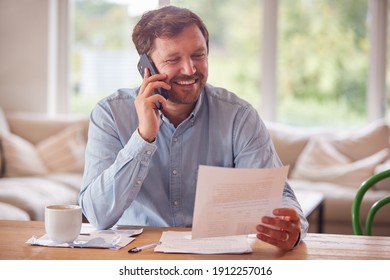 Smiling Man On Phone Call Sitting At Table At Home Reviewing Domestic Finances