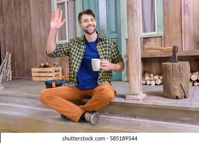 smiling man on porch waving to someone while holding tea cup and looking away