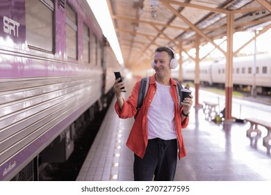 Smiling man on a train platform listens to music with headphones, sips coffee and checks his smartphone while waiting for the next departure, relaxed and connected